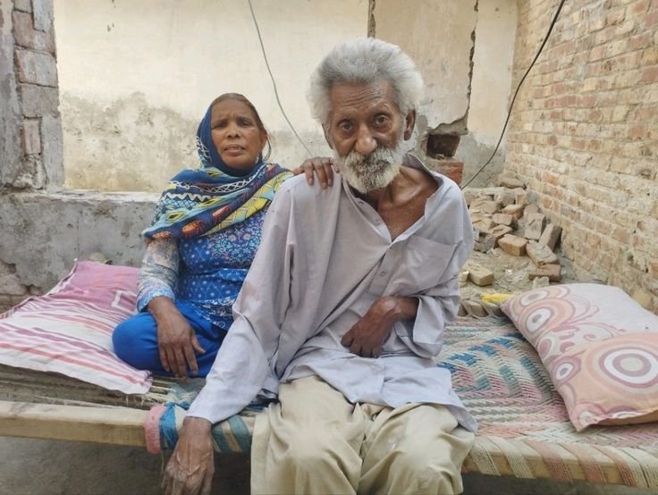 A paralyzed elderly man lying on a bed in a small, poorly maintained house. His wife, also elderly, stands nearby. They are in need of food, medical support, and shelter.